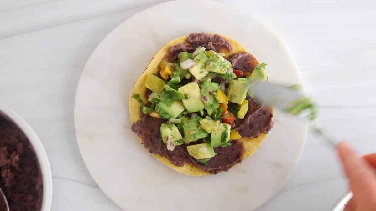 A spoon is topping the refried bean tostadas with the avocado salad.