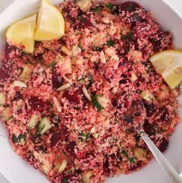 Large white serving bowl with a beet couscous salad topped with lemon wedges and a large serving spoon placed in the salad.