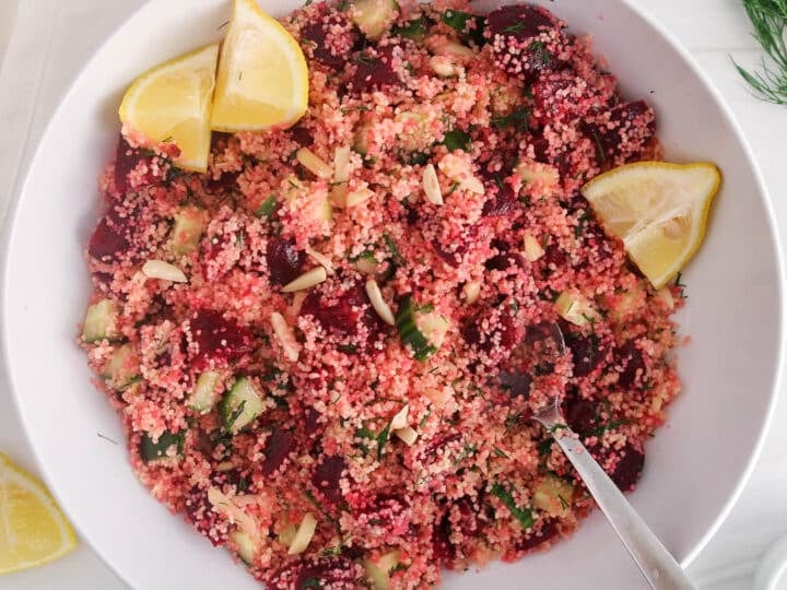 Large white serving bowl with a beet couscous salad topped with lemon wedges and a large serving spoon placed in the salad.