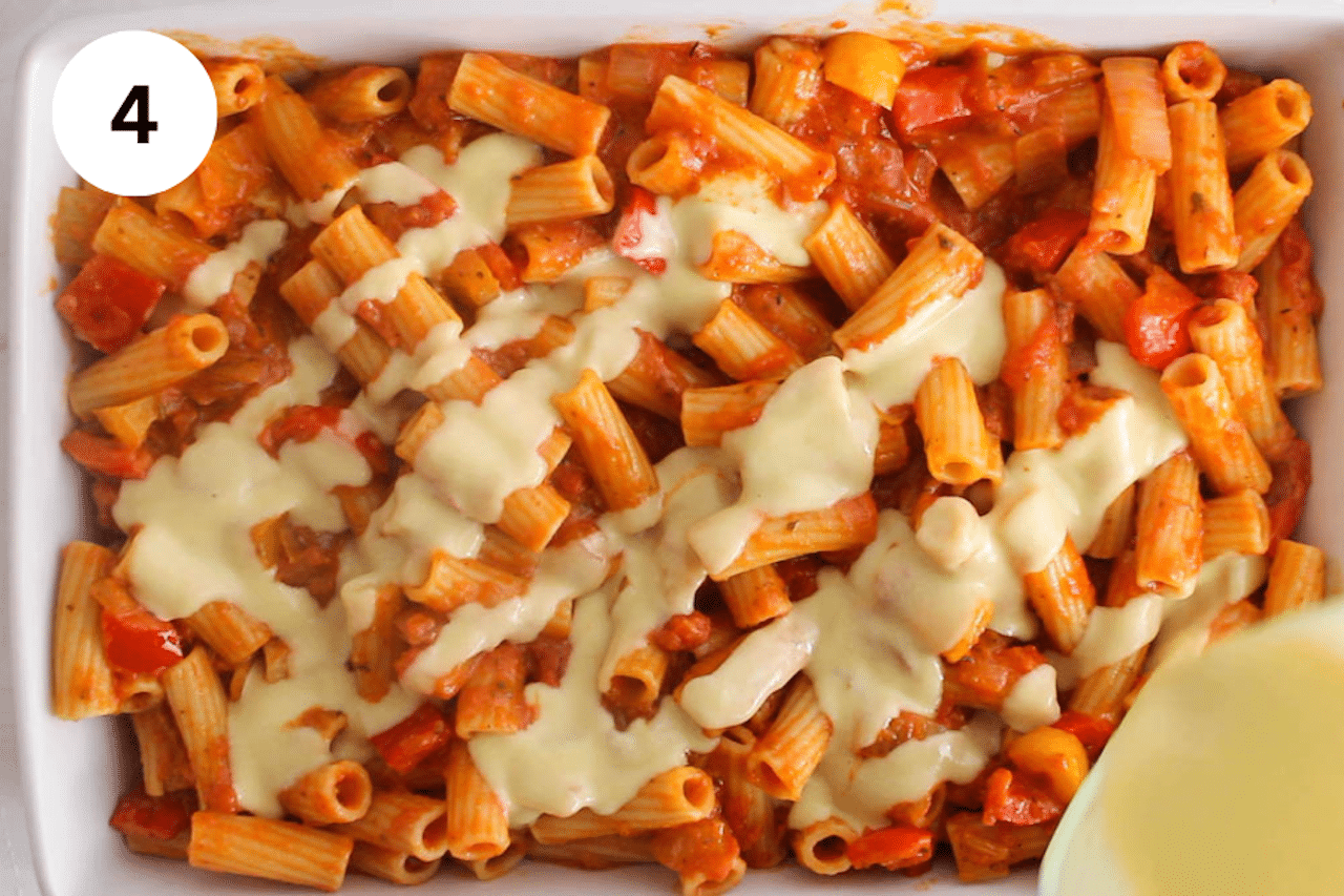 A large baking dish with creamy red sauce pasta while a jar is drizzling a white sauce over.