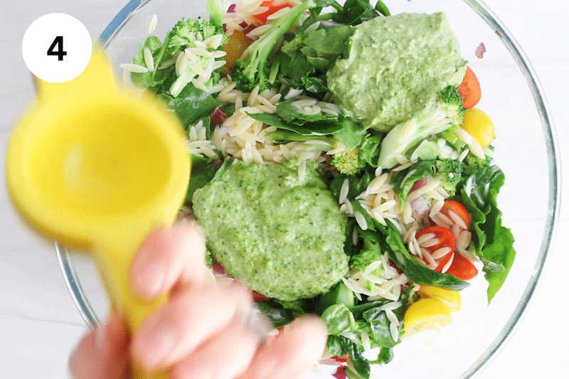A large bowl with veggies and pesto sauce while there are hands squeezing lemon juice over.